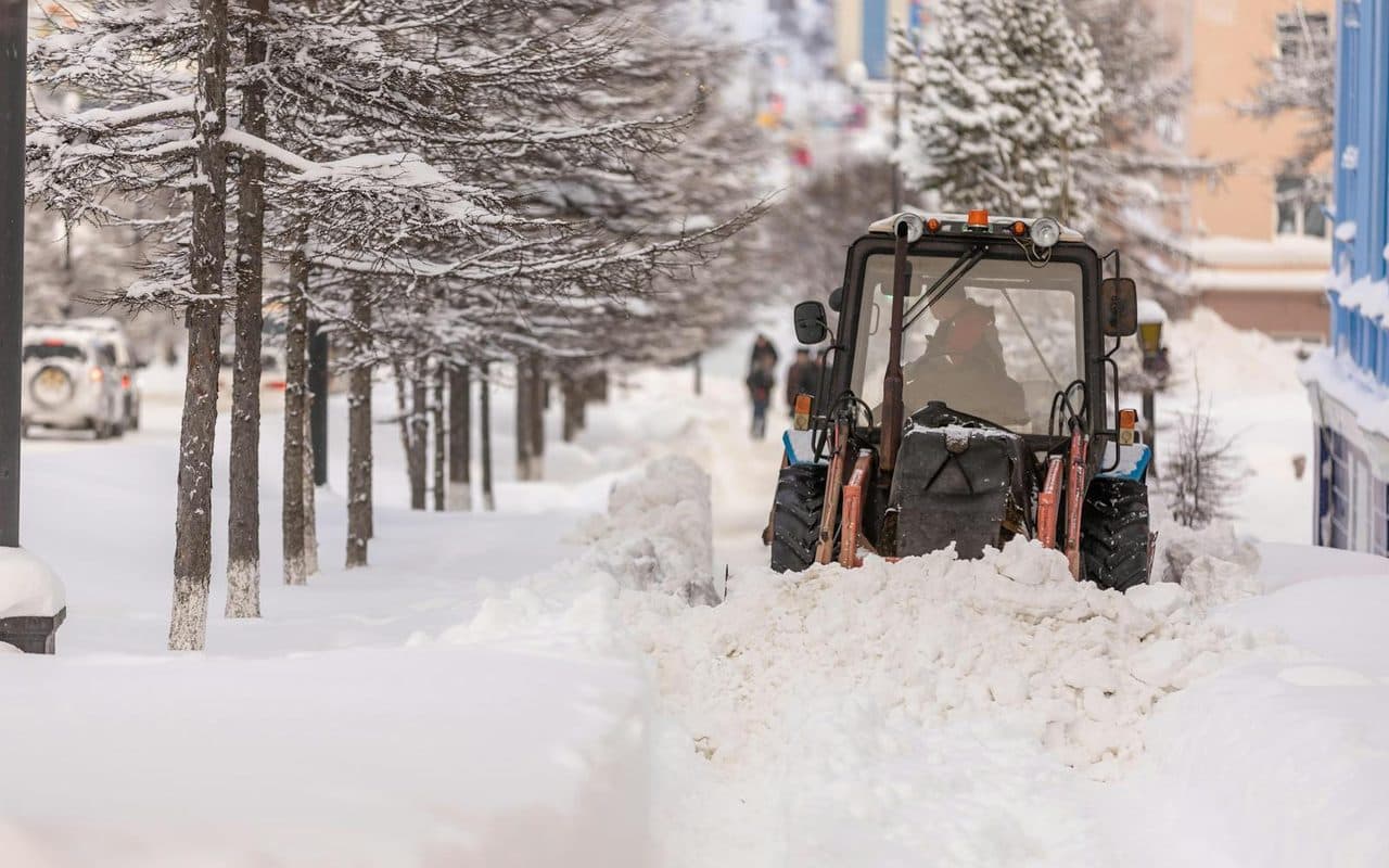 A tractor plow clearing deep snow along a tree-lined commercial route