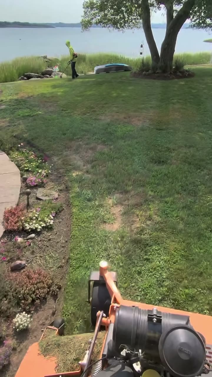 Riding the mower along a waterfront lawn, fresh stripes behind it, a crew member string-trimming the bank