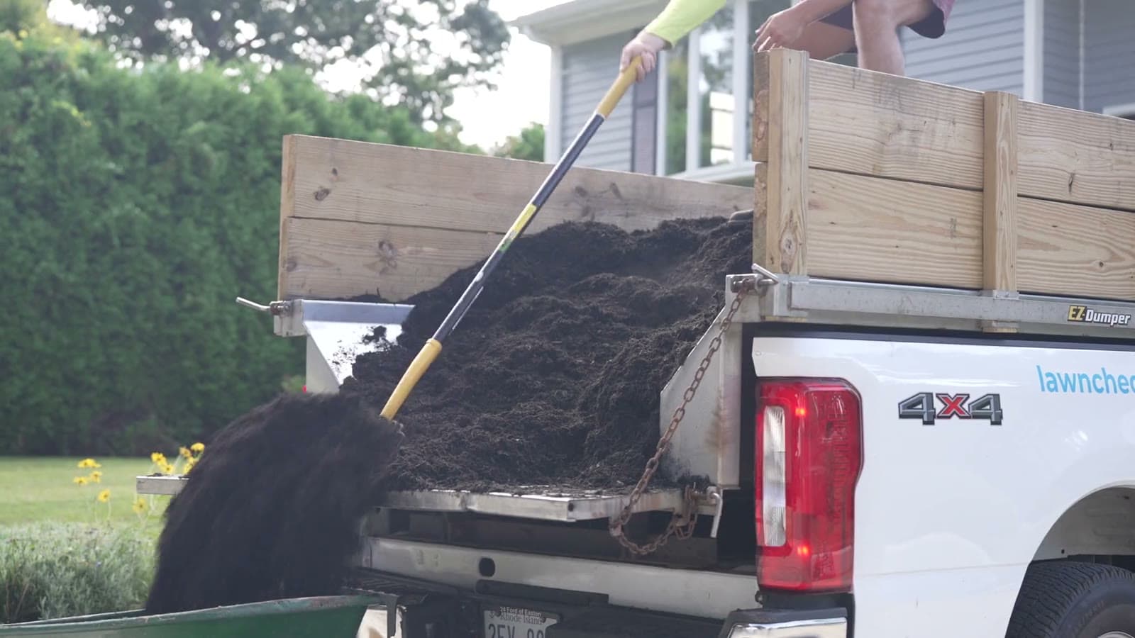 A loaded LawnCheck mulch truck, a full wheelbarrow, a crew member raking a bed and the branded truck on site